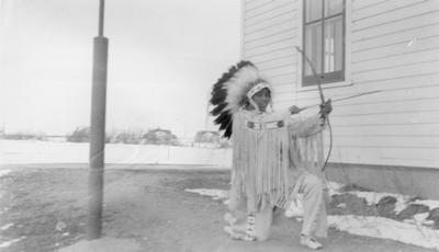 Photo of a young man dressed in ceremonial clothes kneeling and pulling back a bow and arrow, Browning, Montana