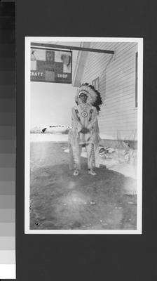 Photo of a boy in ceremonial dress standing outside the Blackfeet Indian Craft Shop, Browning, Montana