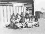 Photo of 12 women and one man sitting in front of a log building sewing, Browning, Montana