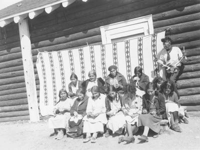 Photo of a group of women and one man sitting in front of a building and sewing, Browning, Montana
