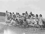 Photo of a large group of women and children sitting outside, some are sewing, Browning, Montana