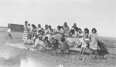 Photo of a large group of women and children sitting outside, some are sewing, Browning, Montana