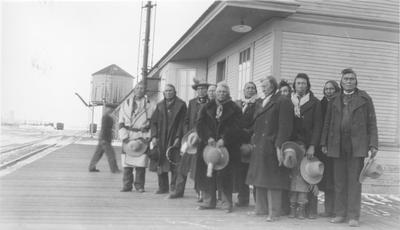 Black and white photo of a large group of Blackfeet men standing in front of the train depot.
