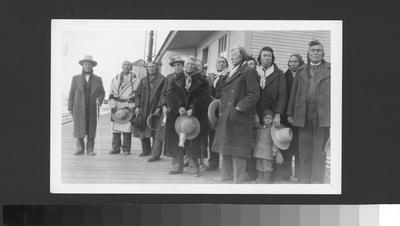Black and white photo of a large group of Blackfeet men and one child standing outside the train depot.