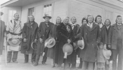 Black and white photo of a group of men, most holding their hats, and children waiting outside the train depot.