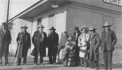 Black and white photo of a group of men and women standing in front of the train depot.