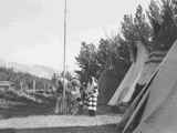 Black and white photo of a group of people in ceremonial dress standing around a flag pole in front of tipis, Saint Mary