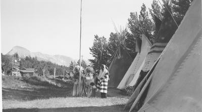 Black and white photo of a group of people in ceremonial dress standing around a flag pole in front of tipis, Saint Mary