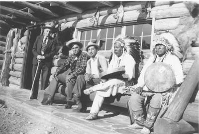 Black and white photo of a group of men, including Apikuni, Lone Wolf, Jim Stingy and Johnny Ground, sitting in front of the craft shop in Stain Mary