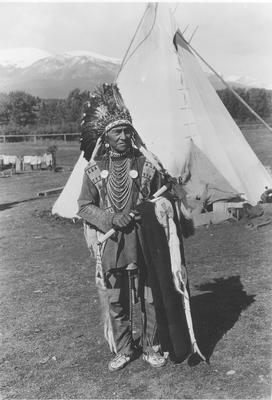 Black and white photo of Wolf Plume in ceremonial dress holding a hatchet and a blanket.