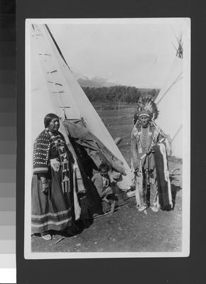 Black and white photo of Wolf Plume, his wife and child, standing in front of a tipi.