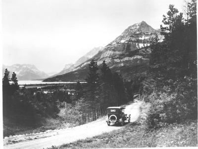 Black and white photo of an automobile driving up a dirt road in Glacier National Park