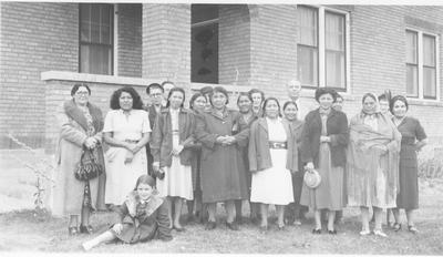 Photo of a large group of women and men standing in front of a brick building.