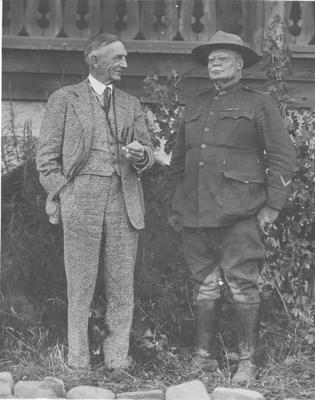 Two Men Standing in Front of a Building, One in a Suit and One in Hunting Clothes, 1919