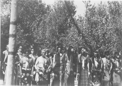 Photo of a group of men including, Bat Weed, Archie Campbell, John LeClair, Eugene Poiree, Ben Perry, Ray Enos and Leon Hobbie, in ceremonial dress standing in front of the center post