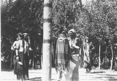 Photo of four men holding feathers face a women with her back to the center post during a ceremony